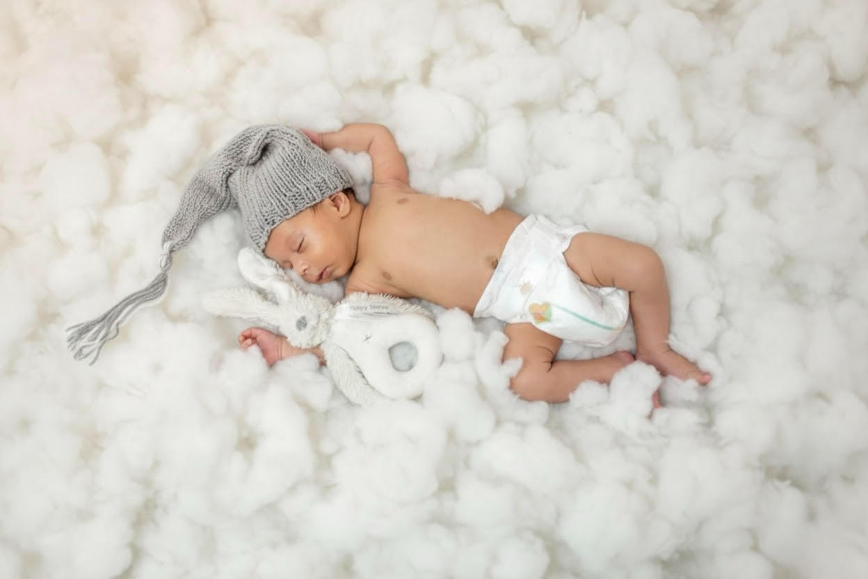 תינוק ישן על מצע רך ובהיר במסגרת ליווי התפתחותי. התינוק חובש כובע ויש לצידו בובה לבנה.